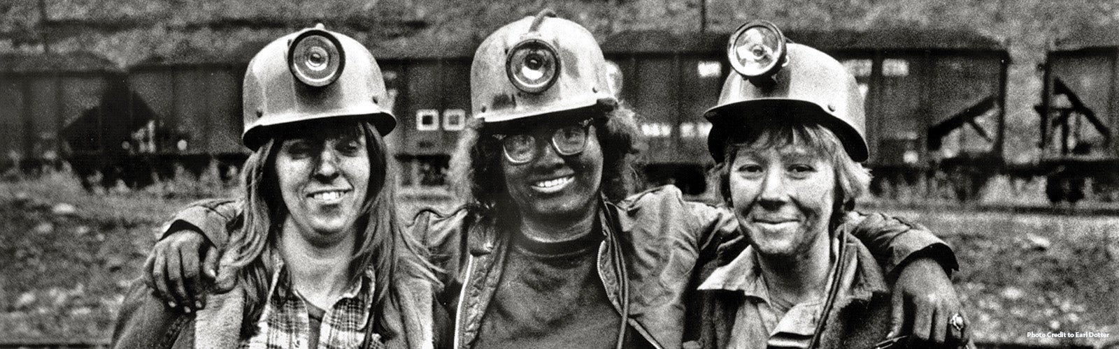 Three female coal miners, wearing helmets with lights, stand arm in arm, smiling. The image is in black and white, with a background of a mining site, including rail cars filled with coal. The miners appear proud and camaraderie is evident among them.