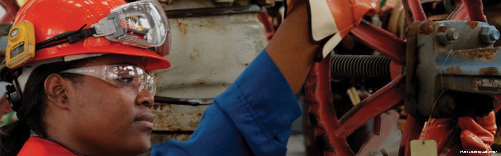 A female steelworker wearing an orange hard hat fitted with goggles and a yellow safety device, along with safety glasses and blue overalls, is operating industrial machinery. The worker's gloved hands are adjusting components on the machine. The background is filled with mechanical parts and equipment.
