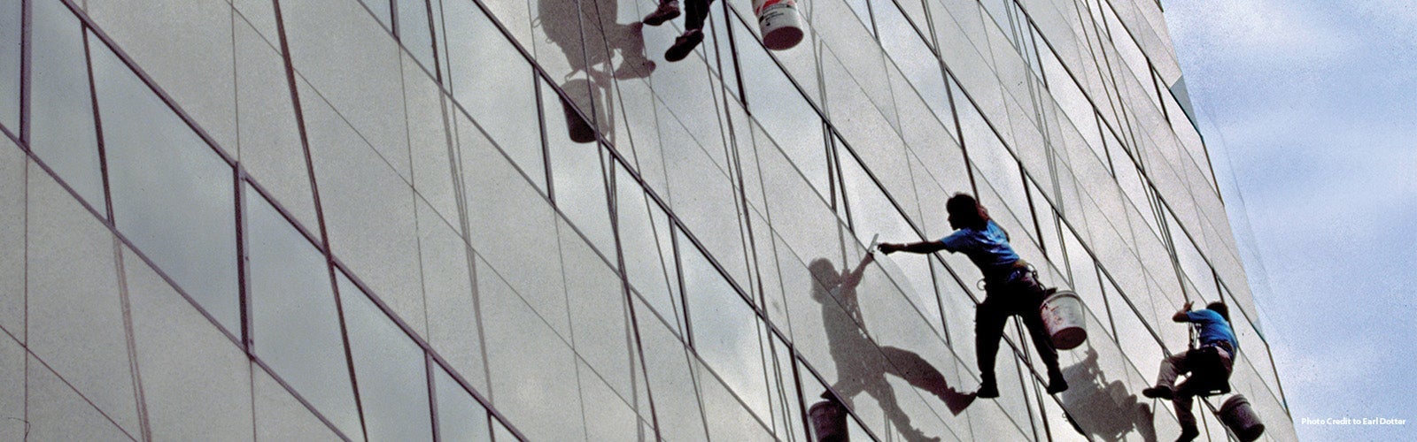 Three window washers are suspended by harnesses as they clean the glass facade of a tall building. Each worker is holding cleaning tools and a bucket, casting shadows on the reflective surface of the windows. The sky is visible on the top right corner of the image.