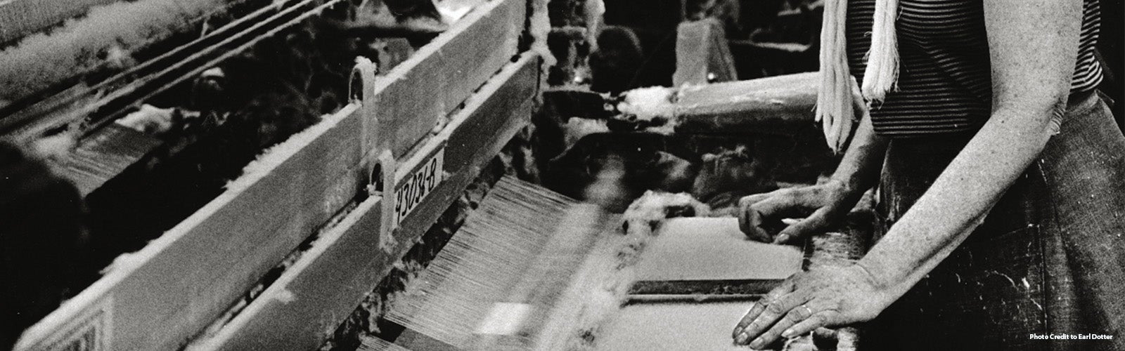 A worker stands at a loom, operating machinery in a textile factory. The image, in black and white, focuses on the worker's hands and the loom's intricate components and threads. The surrounding area appears busy with other machines and indistinct figures, emphasizing the industrious environment. The worker wears a striped shirt and a long scarf-like garment.